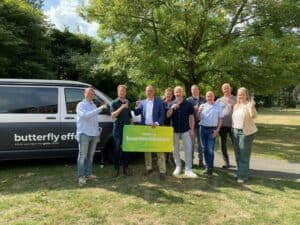 Een groep van acht mensen staat buiten voor een busje met drankjes in de hand en een bord waarop staat: "Welkom buurtverbinders! Vlindereffect.