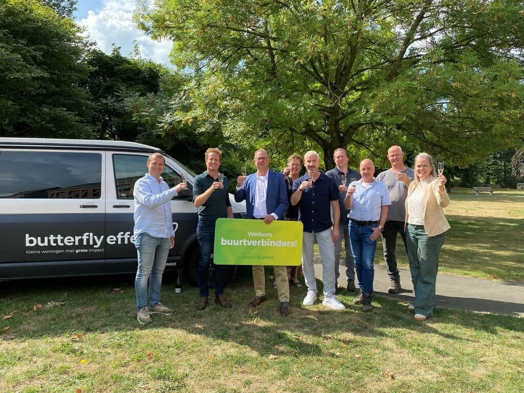 Een groep van acht mensen staat buiten voor een busje met drankjes in de hand en een bord waarop staat: "Welkom buurtverbinders! Vlindereffect.