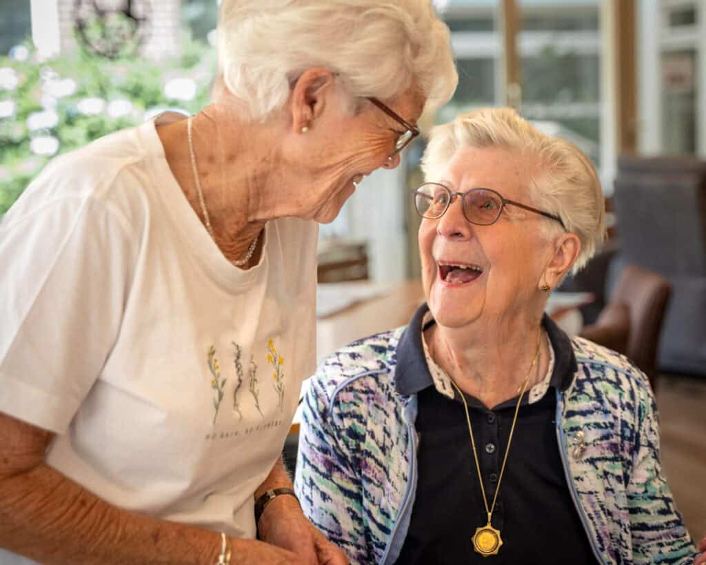 Twee oudere vrouwen binnenshuis, de een staand en lachend naar de ander die zit te lachen, beiden dragen een bril en vrijetijdskleding.