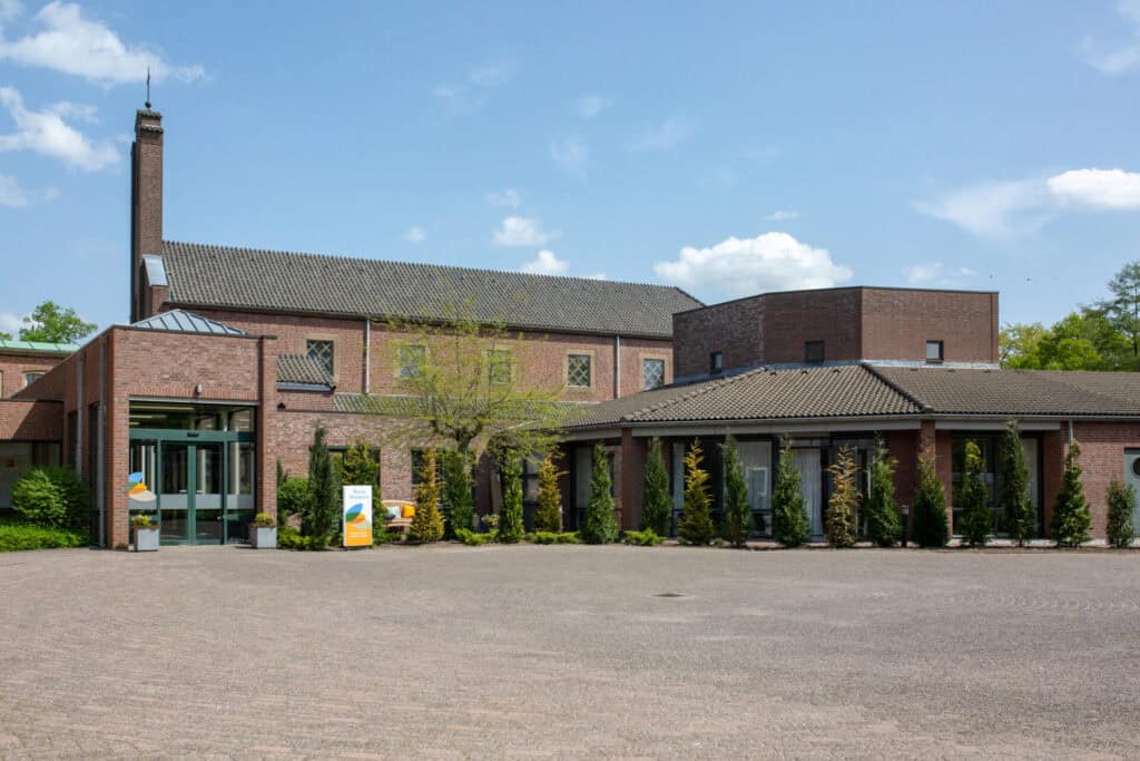Modern bakstenen gebouw met grote ramen, een plat en schuin dak en een hoge schoorsteen, omgeven door groen en een geplaveide binnenplaats onder een strakblauwe hemel.
