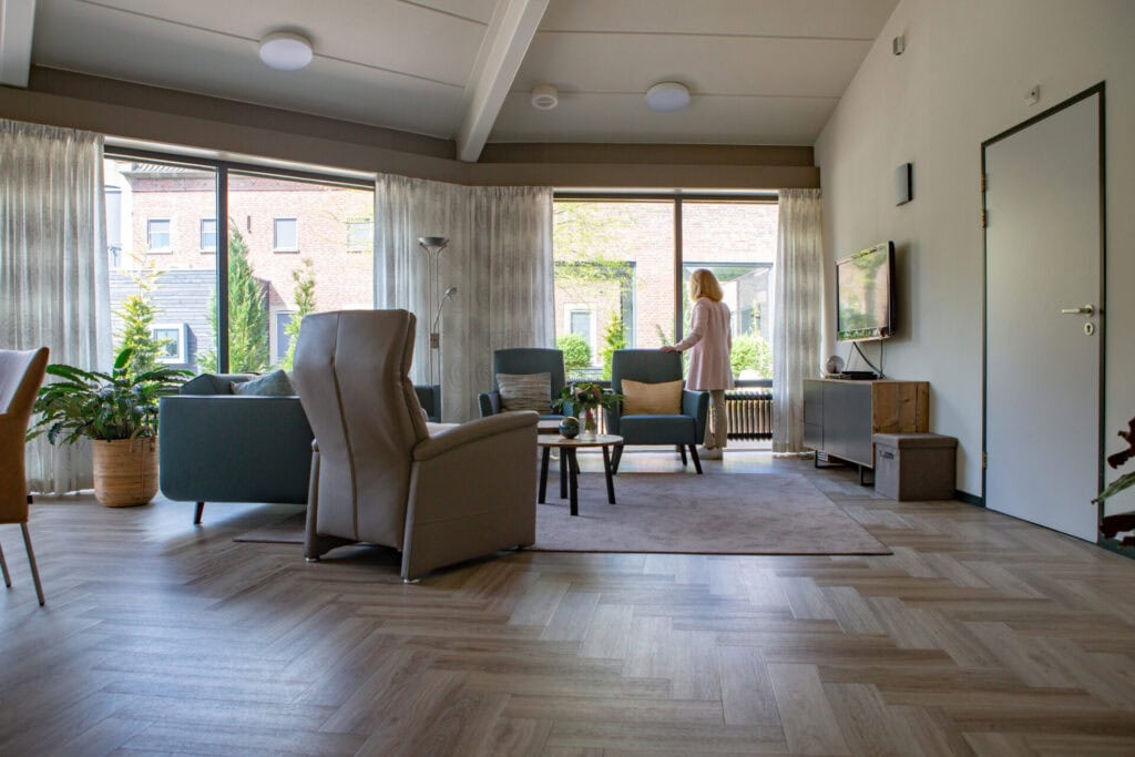 Moderne woonkamer met grote ramen, neutrale meubels, kamerplanten en een vrouw bij het raam die naar buiten kijkt.