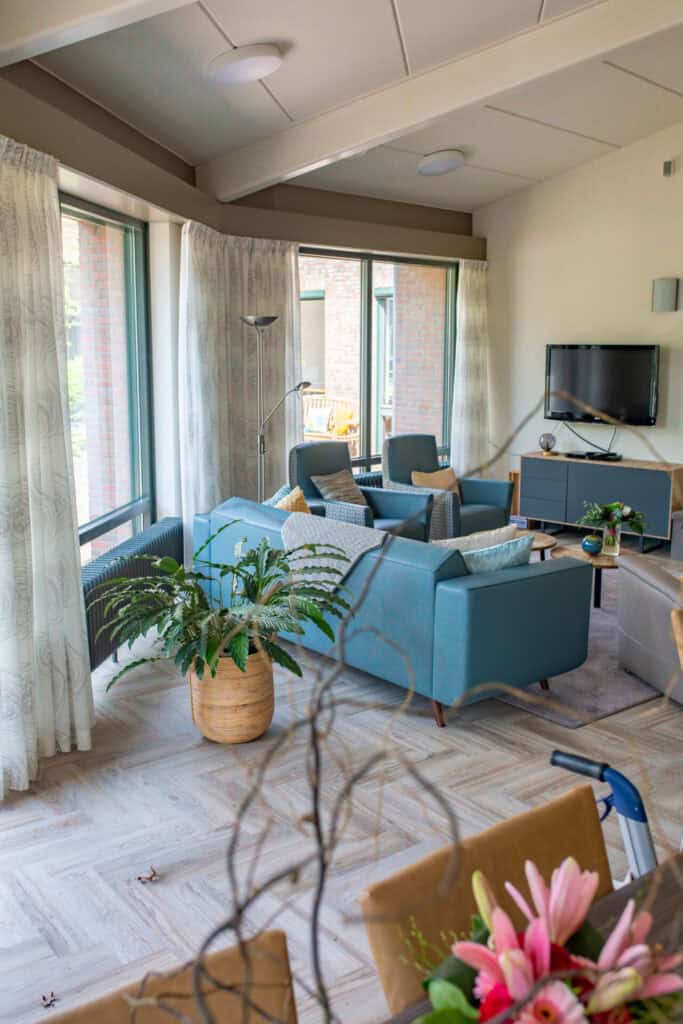 Moderne woonkamer met grote ramen, blauw bankstel, potplanten, een tv aan de muur en lichte houten vloeren.