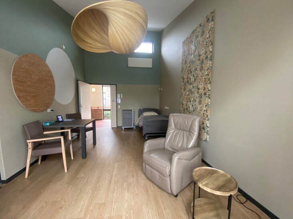 Een moderne, minimalistische kamer met een eenpersoonsbed, een fauteuil, een tafel met twee stoelen, een bijzettafel en decoratieve muurelementen onder grote hanglampen.