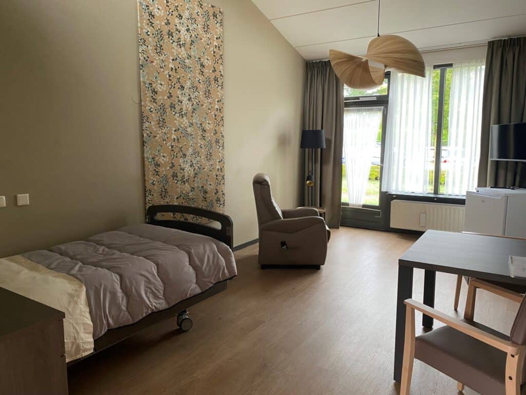 Een eenvoudige, moderne slaapkamer met een eenpersoonsbed, een relaxstoel, een bureau met stoelen, een groot raam met lichte gordijnen en een staande lamp.