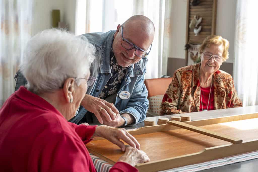 Twee oudere vrouwen en een man spelen samen een houten bordspel aan een tafel, lachend en genietend van de activiteit in een lichte kamer.