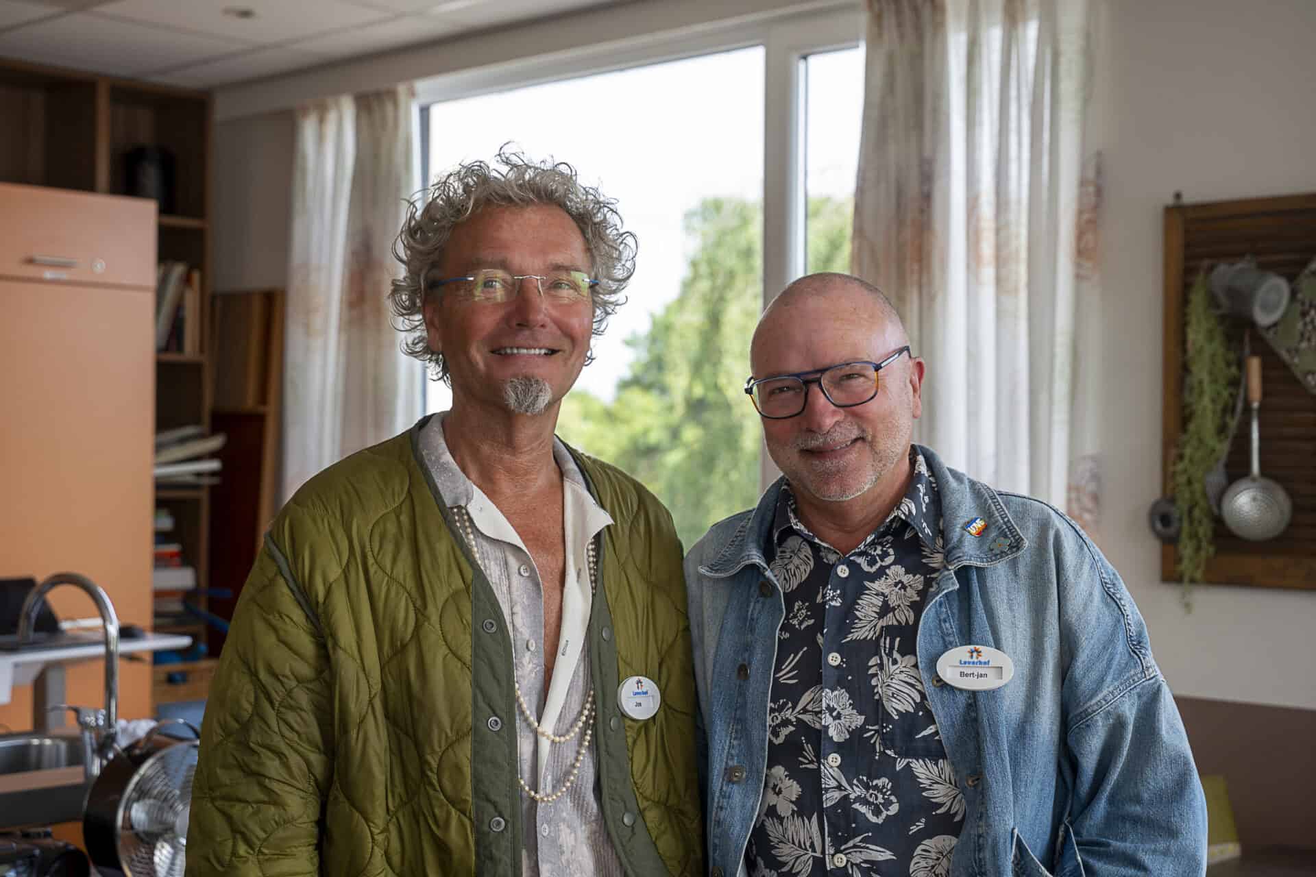 Twee mannen staan binnen naast elkaar en glimlachen naar de camera. Ze dragen naamplaatjes en vrijetijdskleding en er is een raam met gordijnen op de achtergrond.