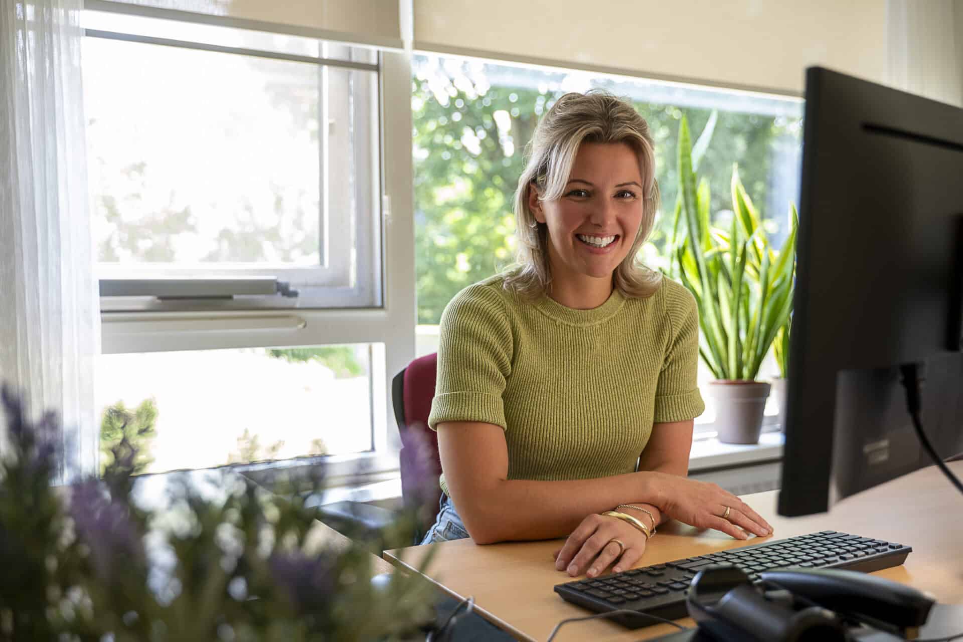 Een vrouw zit aan een bureau voor een computerscherm en lacht naar de camera. Op de achtergrond staan planten en een raam met natuurlijk licht.