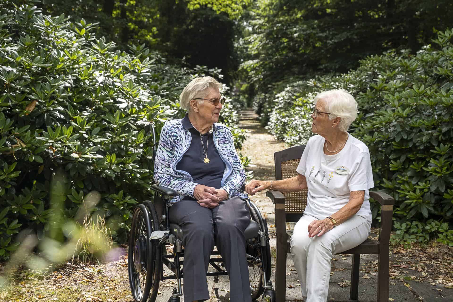 Twee oudere vrouwen, één in een rolstoel en één op een bankje, zitten buiten op een tuinpad omgeven door groen en zijn met elkaar in gesprek.