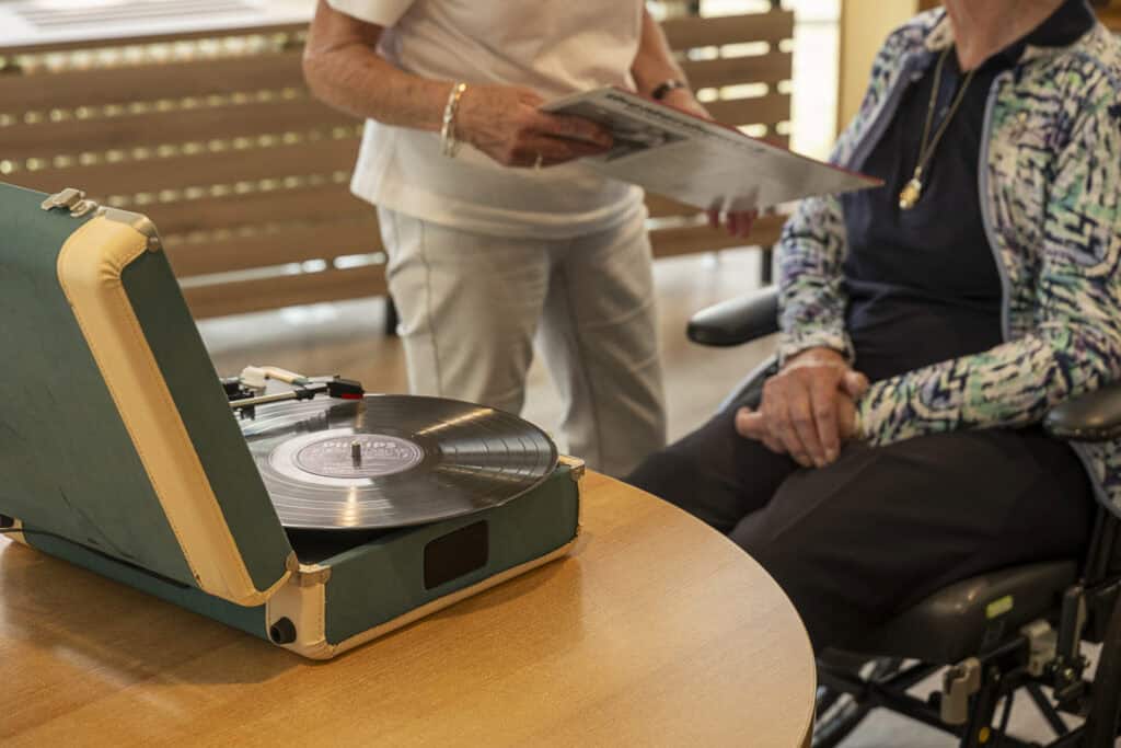 Twee mensen staan en zitten bij een tafel met een vinylplaat die wordt afgespeeld op een draagbare platenspeler; één persoon houdt een platenhoes vast.