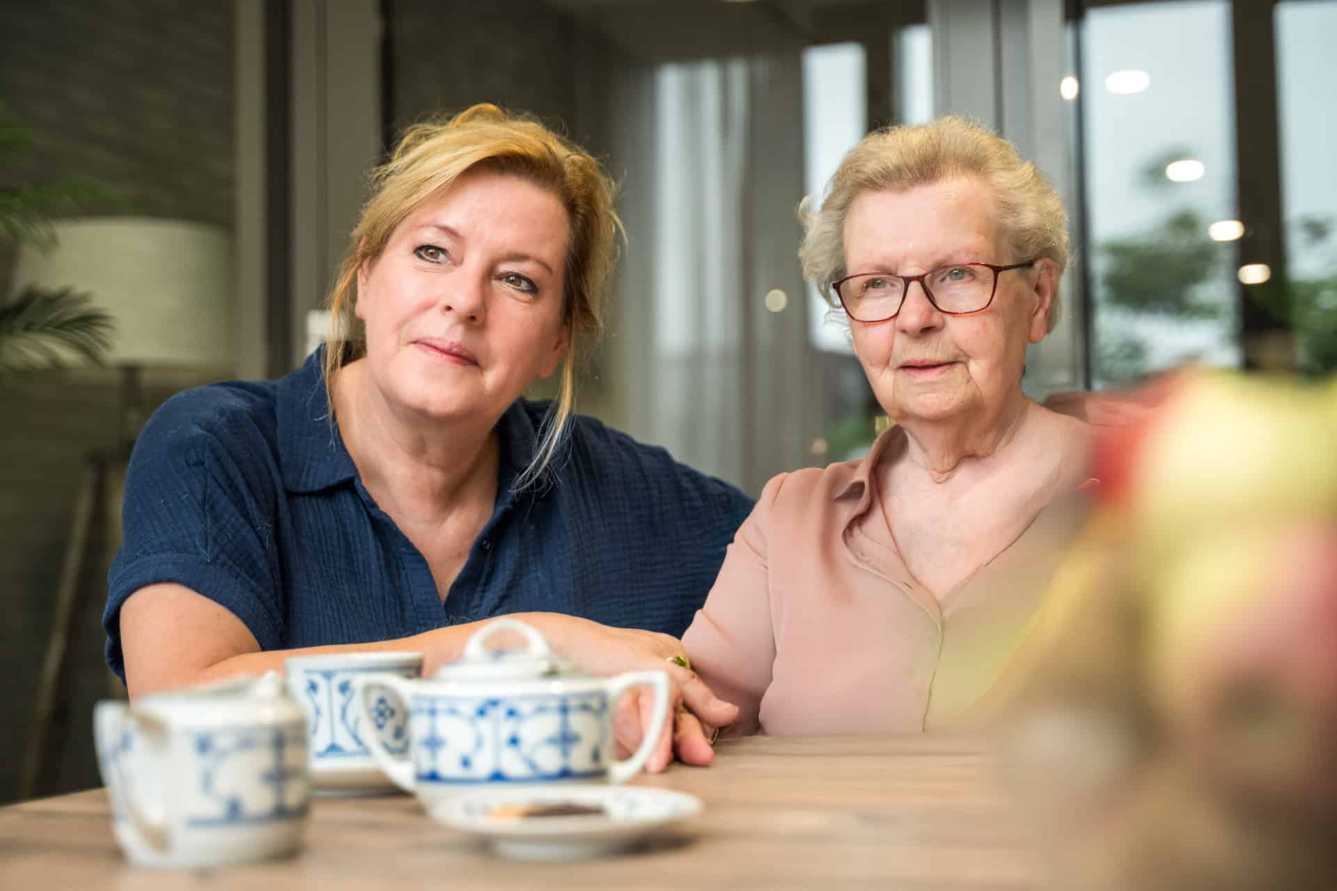 Twee vrouwen, een jongere en een oudere, zitten samen aan een tafel met theekopjes en schoteltjes en kijken in de verte in een lichte, overdekte omgeving.