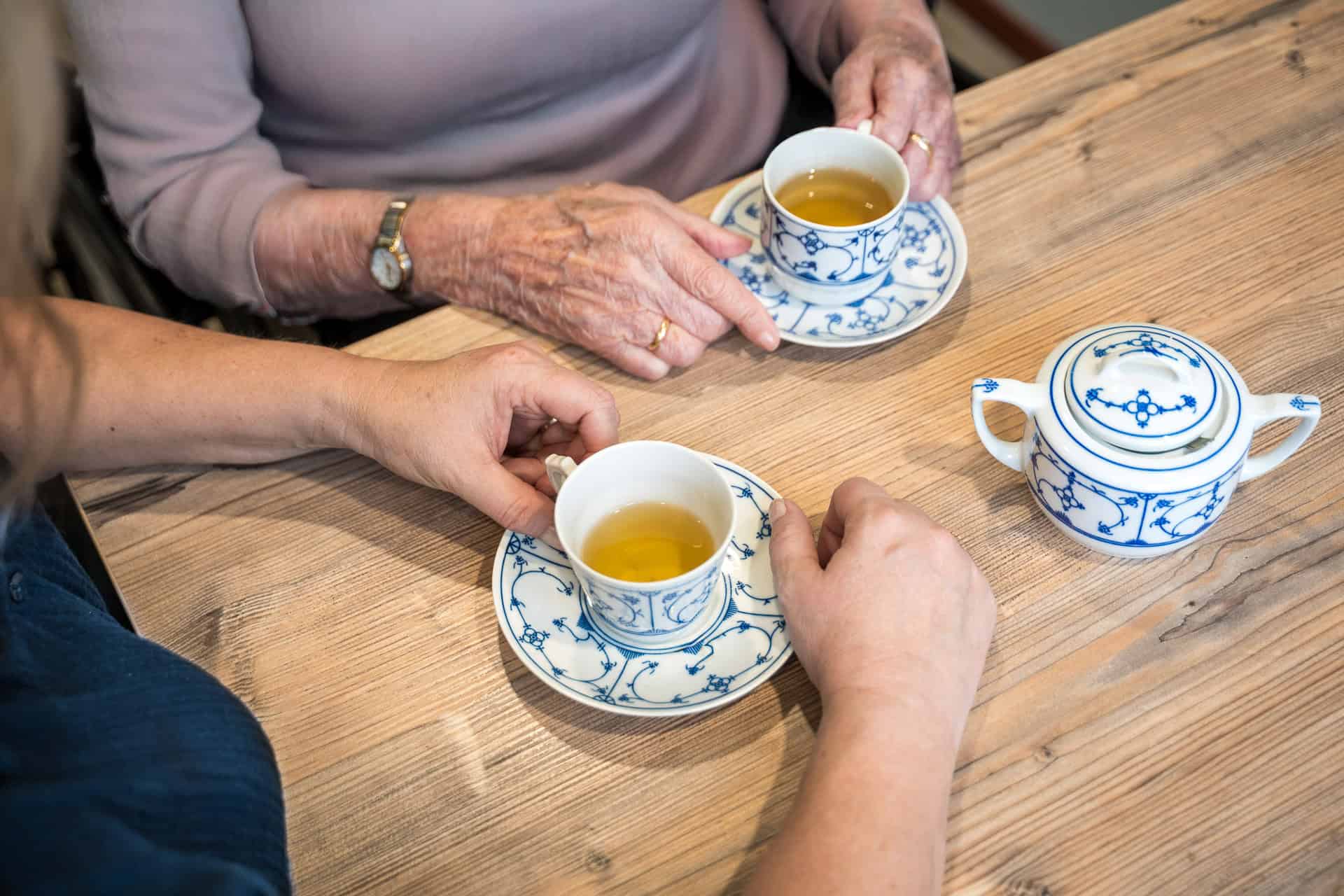 Twee mensen zitten aan een houten tafel, elk met een kopje thee in blauw en wit porselein, met een bijpassende suikerpot naast zich.