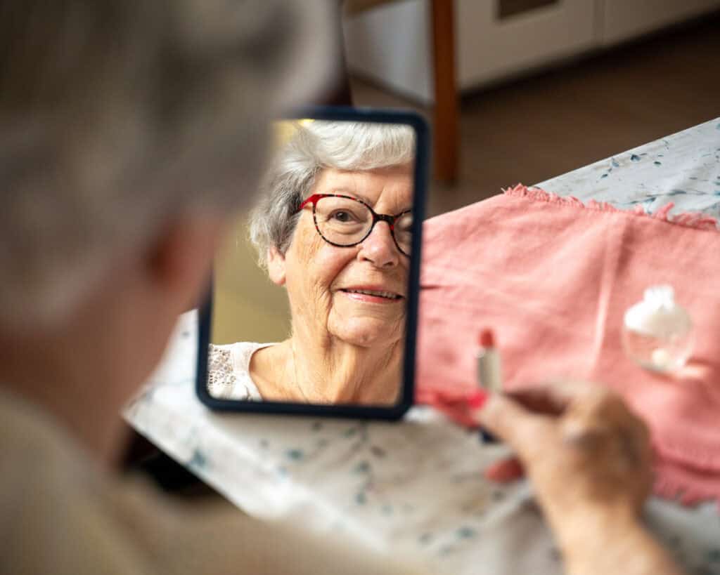 Een oudere vrouw met bril kijkt naar haar spiegelbeeld terwijl ze een tube lippenstift vasthoudt aan een tafel met een roze doek en een klein potje.