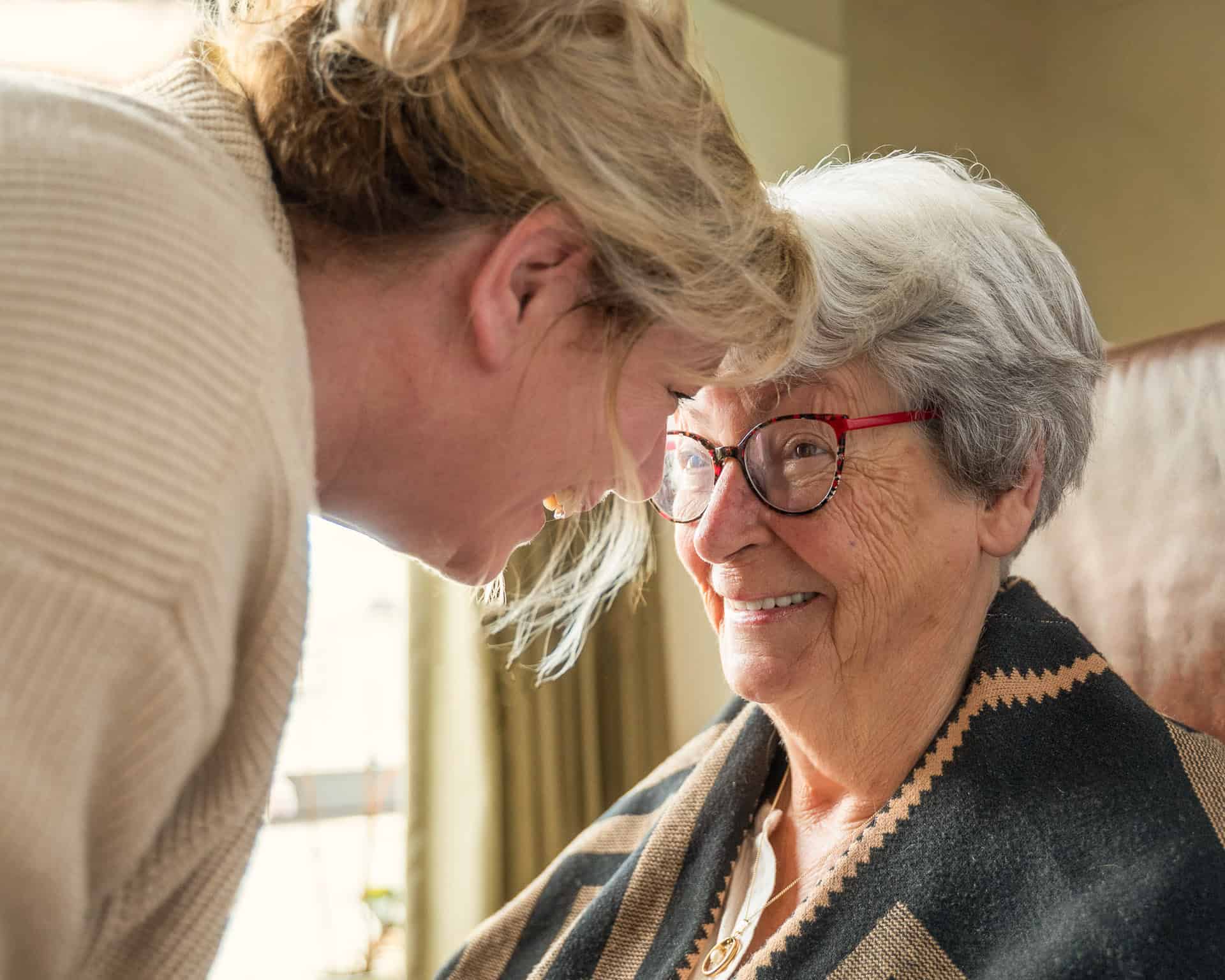 Een oudere vrouw met een bril glimlacht als een jongere vrouw naar haar toe leunt en haar voorhoofd aanraakt in een warme, zonovergoten kamer.