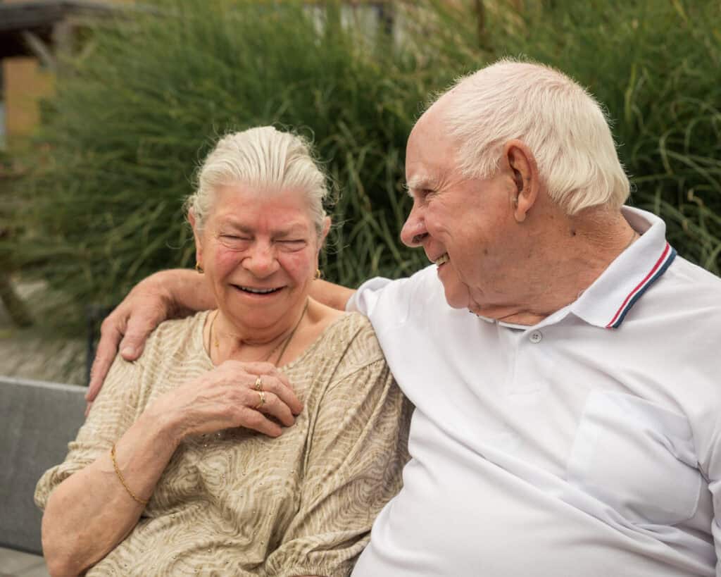 Een oudere man en vrouw zitten dicht tegen elkaar aan op een bankje buiten, lachend en glimlachend, met groen op de achtergrond.