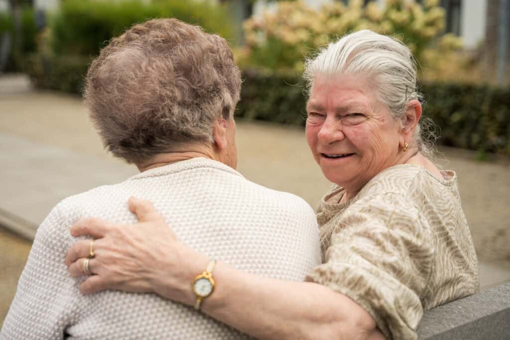 Twee oudere vrouwen zitten buiten op een bankje, de een met haar arm om de ander heen. De ene vrouw kijkt met haar gezicht naar de camera en glimlacht, terwijl de andere met haar rug naar haar toe staat.