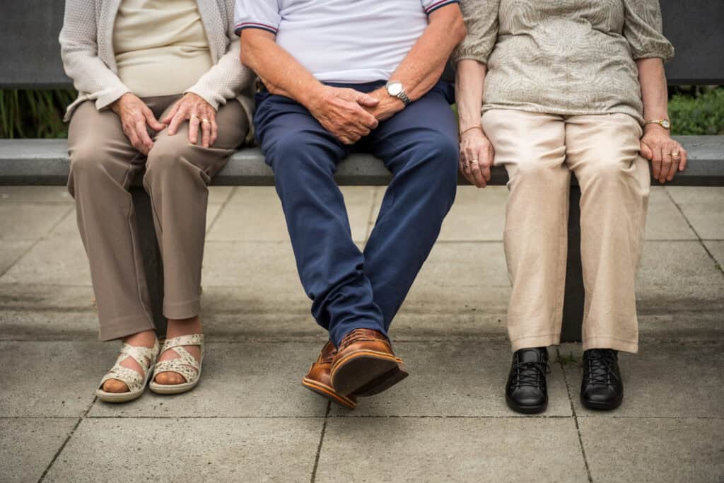 Drie ouderen zitten buiten naast elkaar op een bankje, met hun handen op hun schoot en alleen hun onderlichaam en benen zichtbaar.