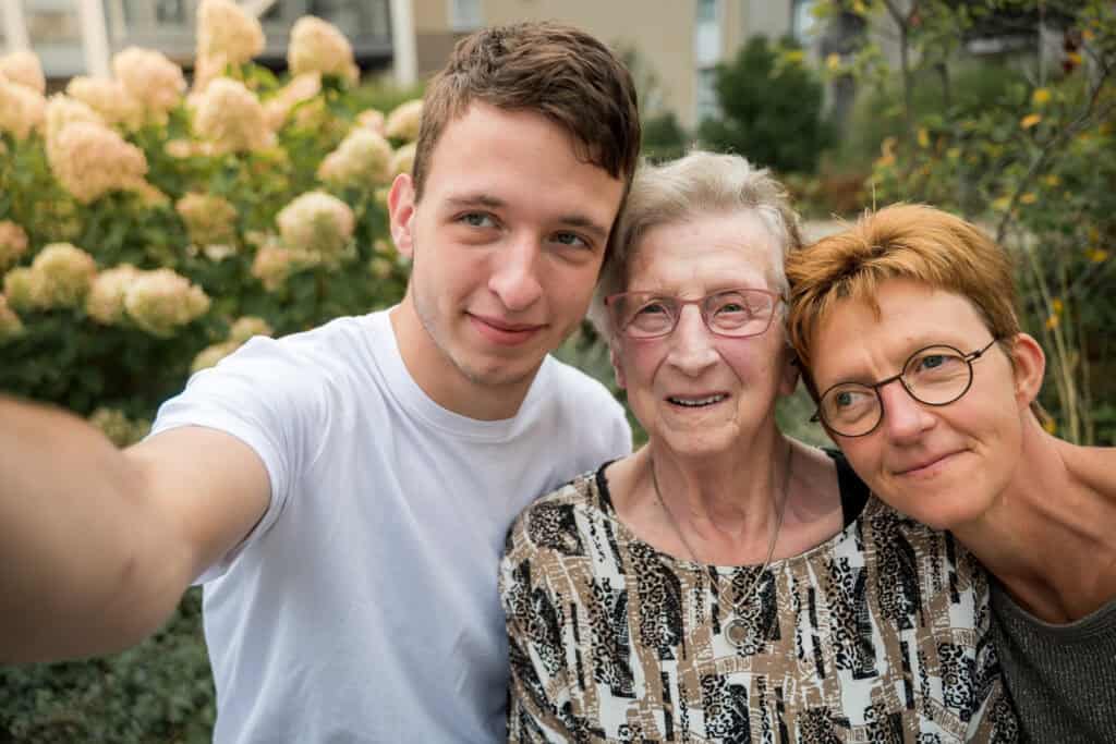 Drie mensen, een jonge man, een oudere vrouw en een vrouw van middelbare leeftijd, staan buiten dicht bij elkaar en nemen een selfie met bloeiende hortensia's op de achtergrond.