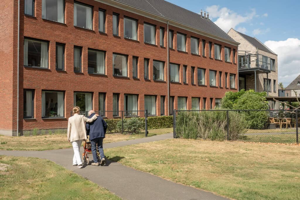 Twee mensen, van wie er één een rollator gebruikt, lopen op een zonnige dag over een pad buiten een stenen flatgebouw of zorginstelling.
