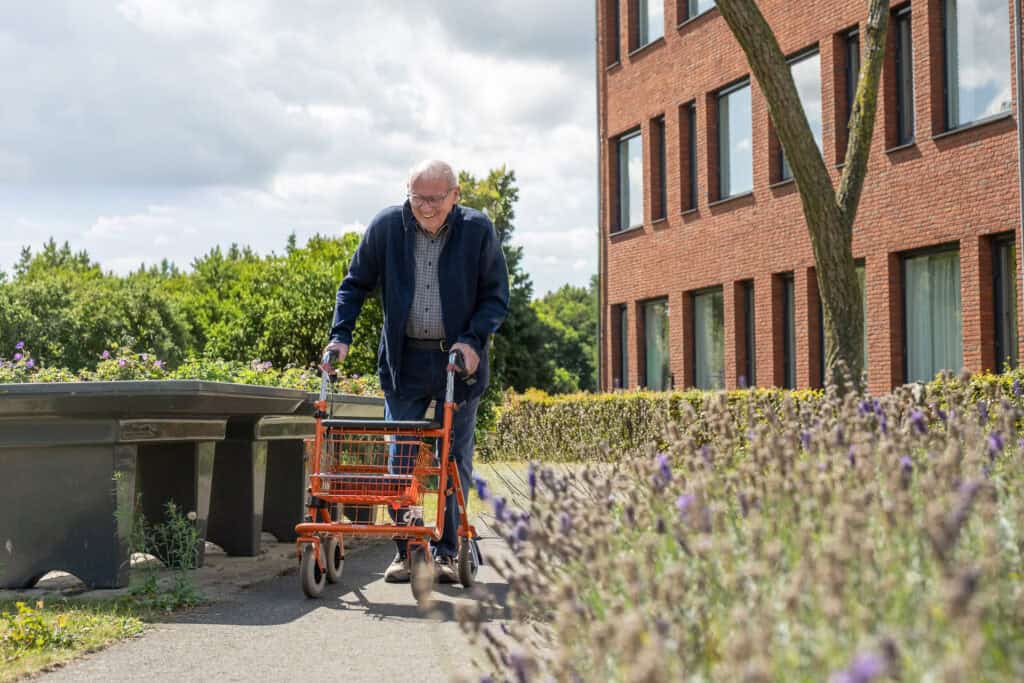 Een oudere man gebruikt een rollator op wielen terwijl hij over een tuinpad naast een stenen gebouw loopt op een bewolkte dag.