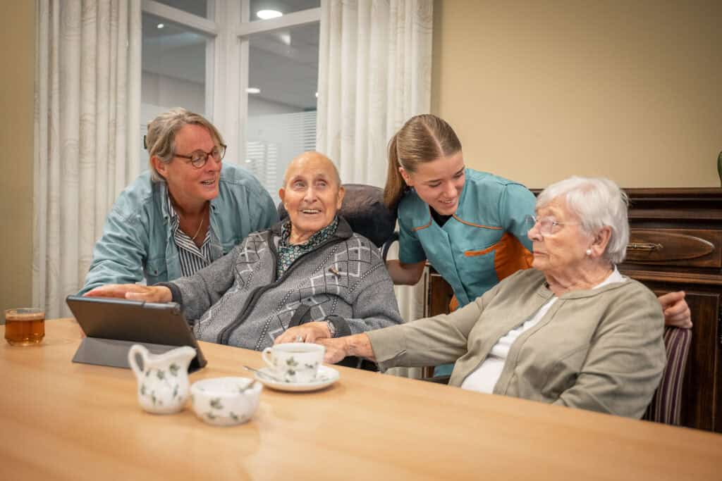 Twee ouderen zitten aan een tafel met theekopjes, samen met twee verzorgers; één persoon gebruikt een tablet terwijl de groep contact heeft in een lichte, comfortabele kamer.