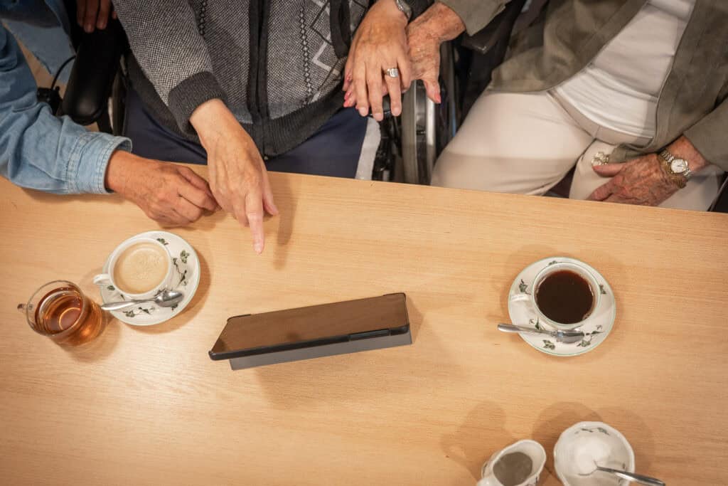 Drie ouderen zitten aan een tafel met koffie en thee, één persoon wijst naar een tablet in het midden.