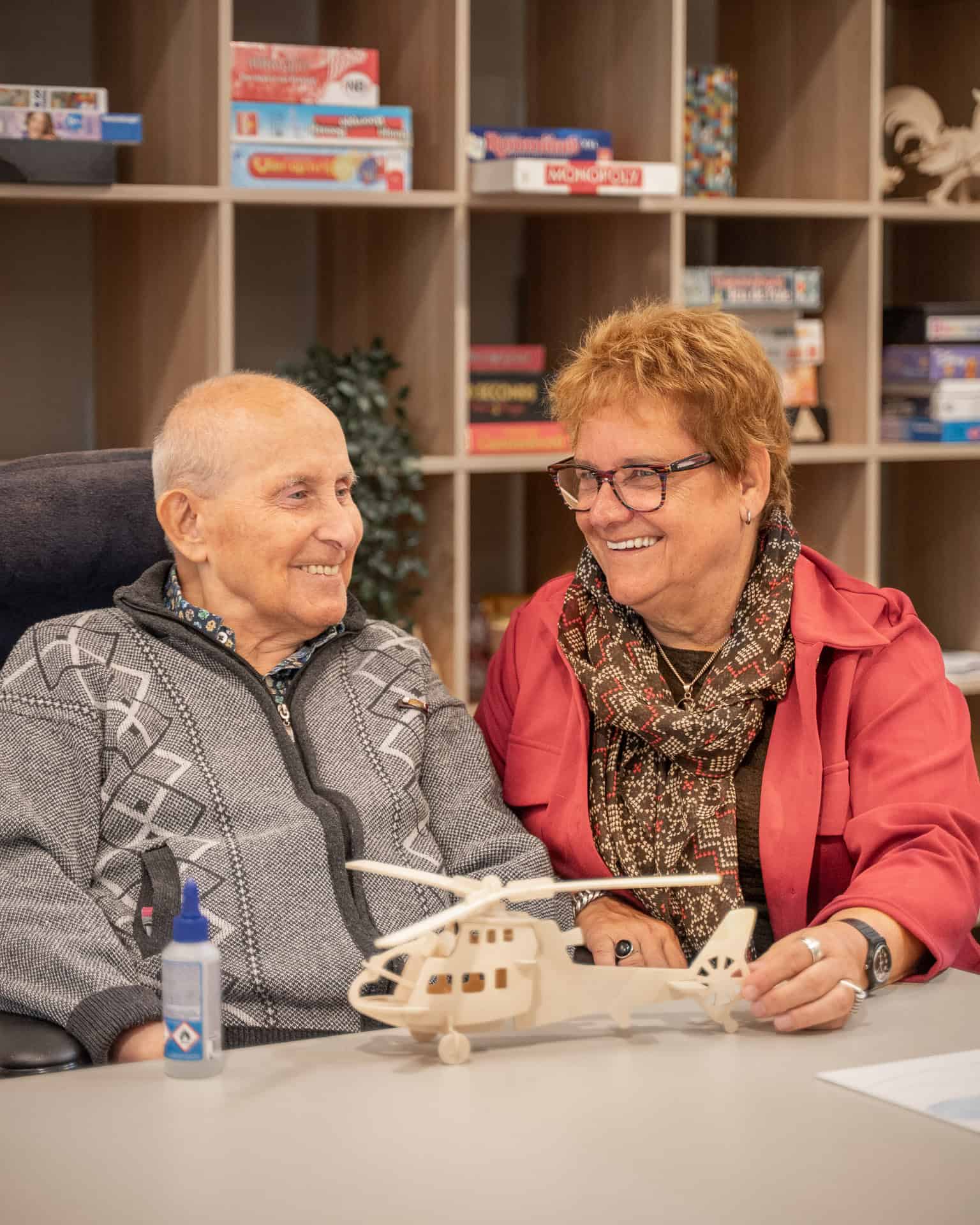 Een oudere man en vrouw zitten glimlachend aan een tafel, houden een houten modelhelikopter vast, met bordspellen op planken op de achtergrond.