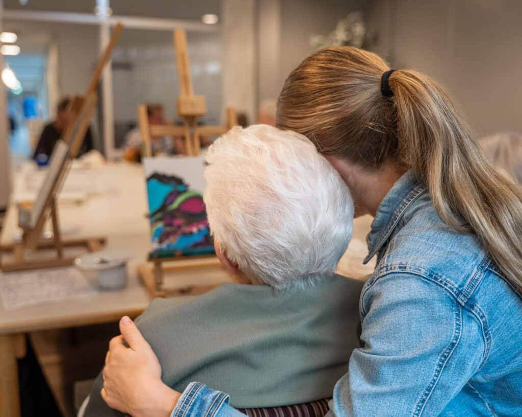 Een jongere vrouw omhelst een oudere vrouw van achteren terwijl ze kijken naar een kleurrijk schilderij op een ezel in een kunstklasomgeving.