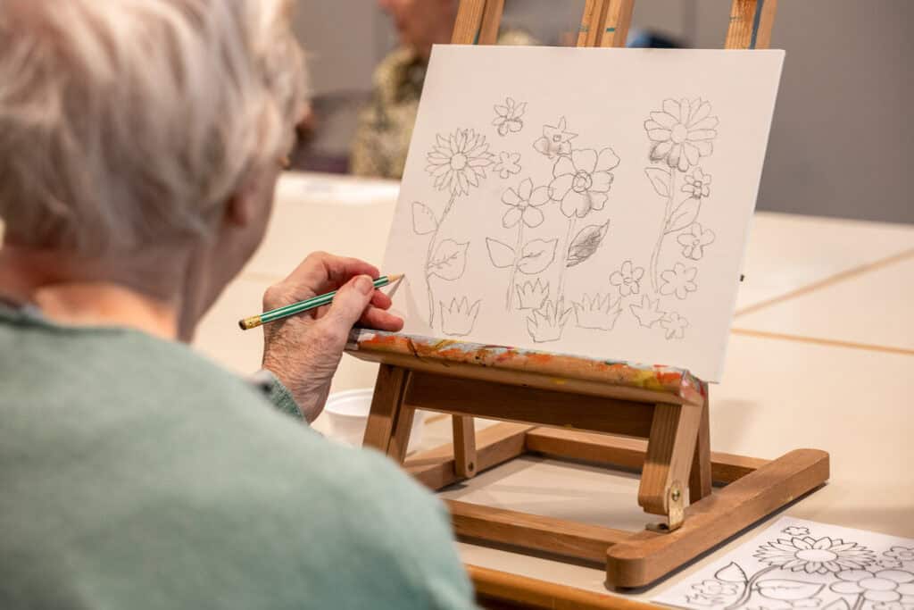 Een oudere persoon schetst bloemen in potlood op een doek aan een ezel, met een andere bloementekening zichtbaar op de tafel vlakbij.