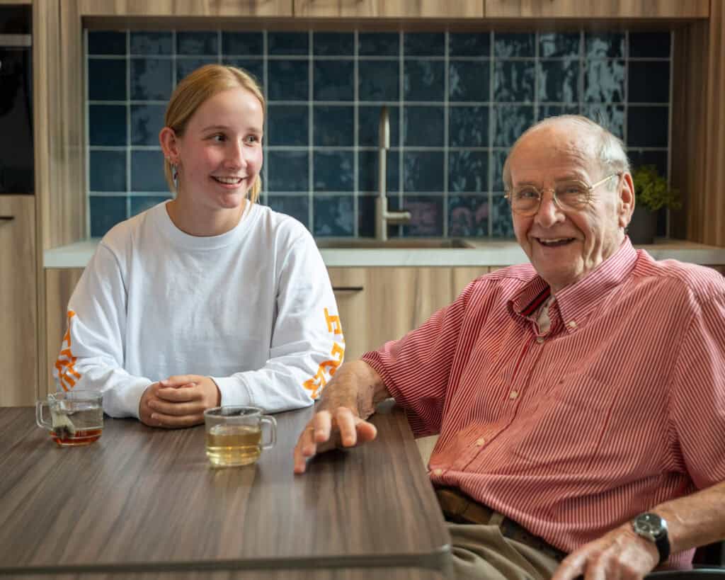 Een jonge vrouw en een oudere man zitten aan een tafel met twee kopjes thee, glimlachen en praten in een moderne keuken.