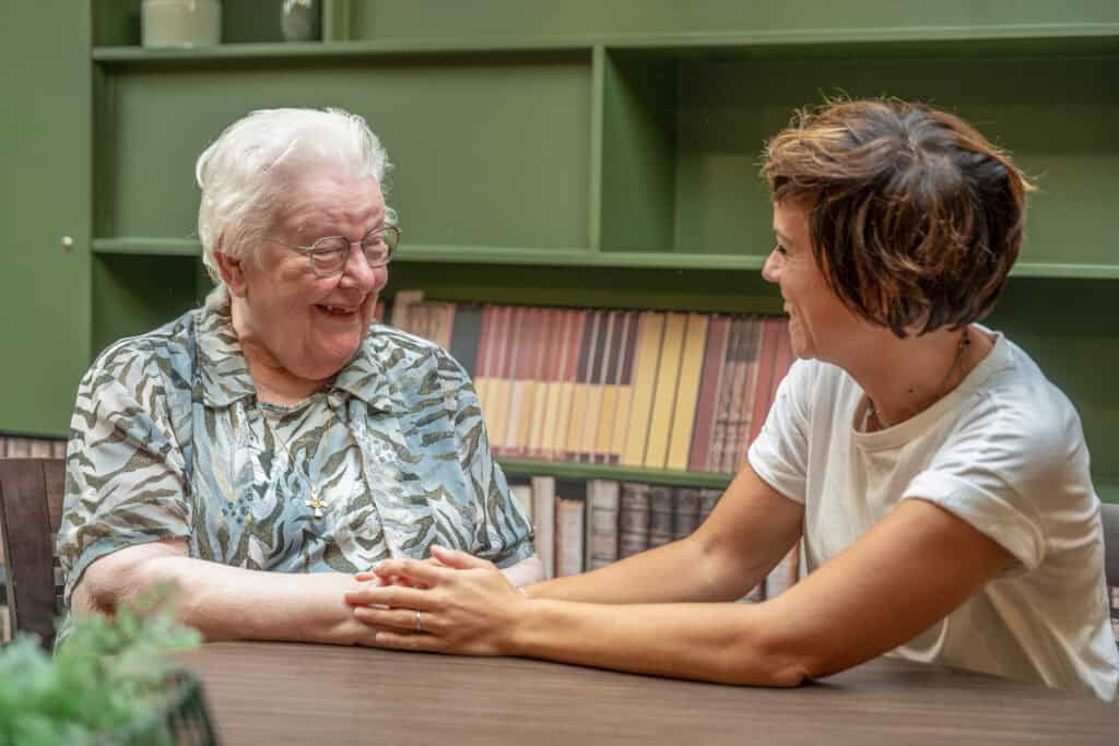 Een oudere vrouw en een jongere vrouw zitten binnen aan een tafel, glimlachen en houden elkaars hand vast terwijl ze een gesprek voeren.