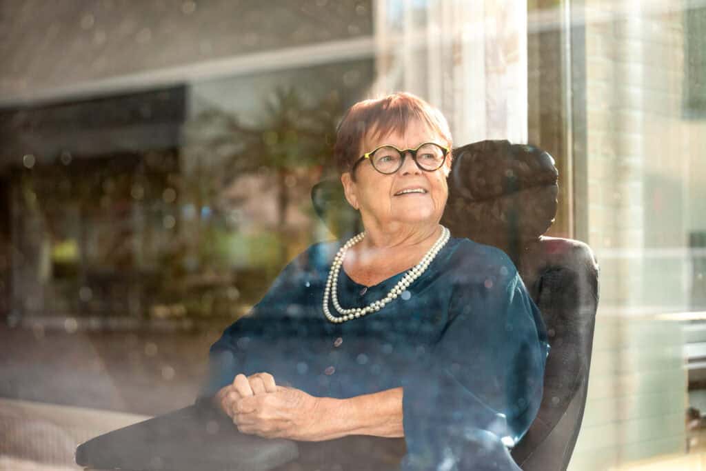 Een oudere vrouw met kort haar en een bril zit in een stoel, glimlacht en kijkt door een raam naar buiten.