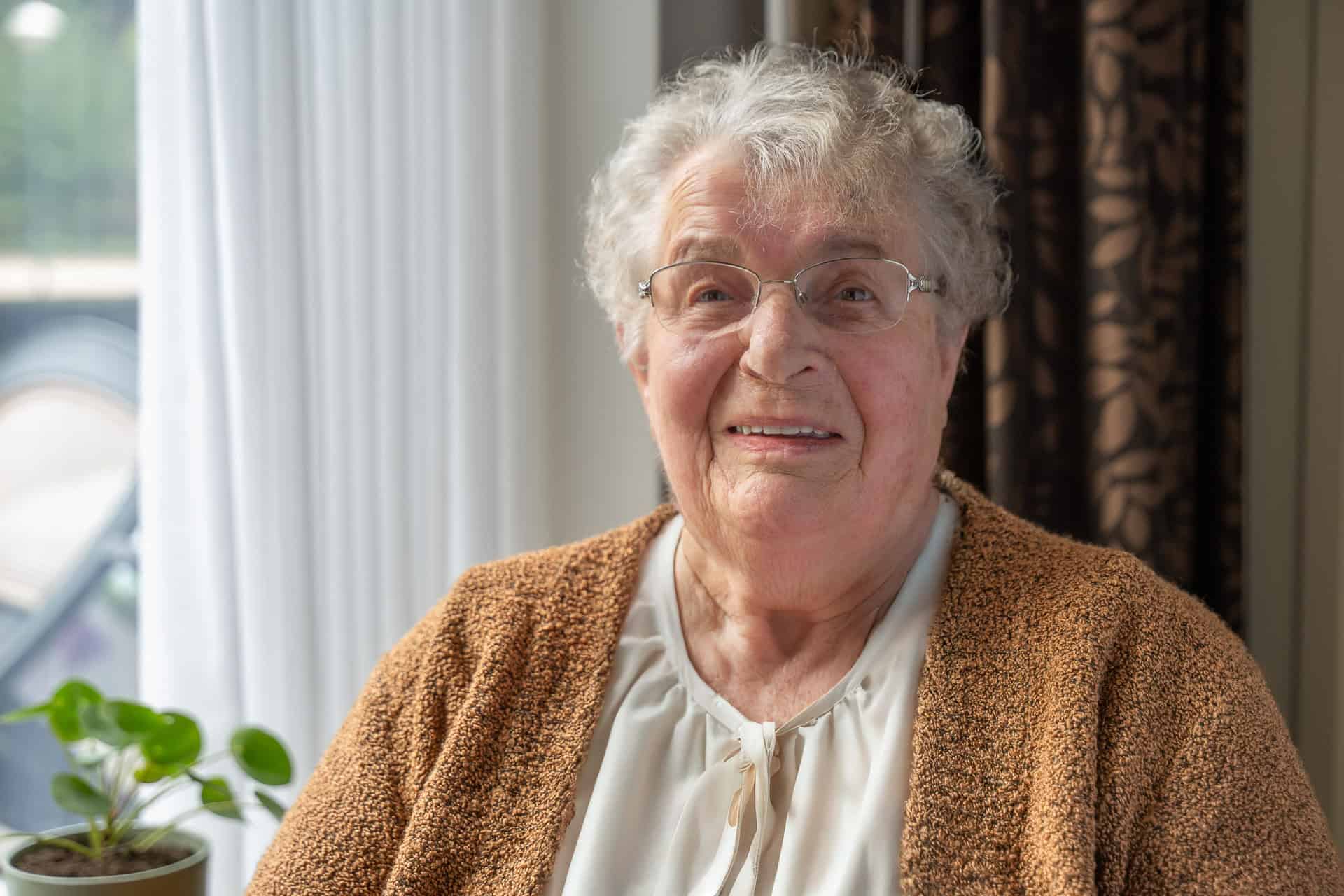 Een oudere vrouw met grijs haar, een bril en een bruin vest zit binnen bij een raam met een potplant naast haar.