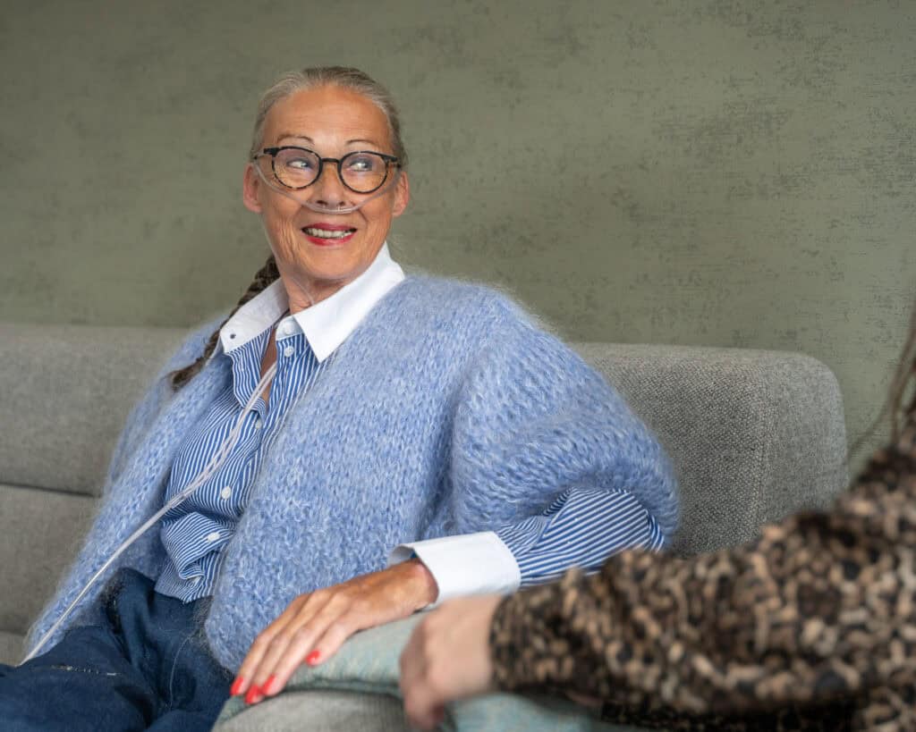 Oudere vrouw met bril en neuscanule zit op een grijze sofa, glimlacht en praat met een andere persoon wiens arm zichtbaar is op de voorgrond.