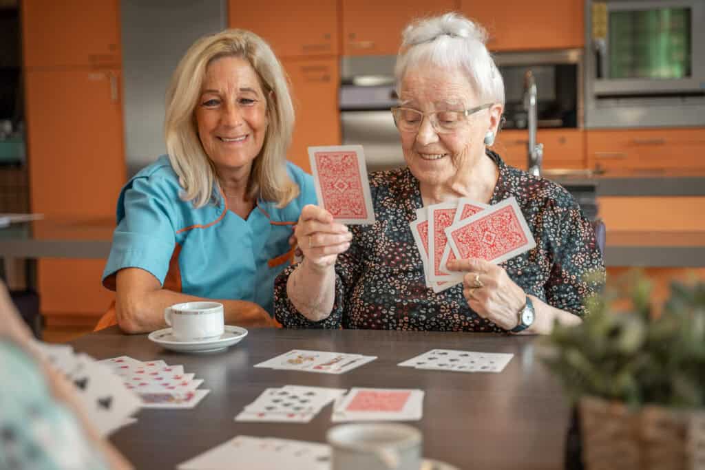 Een oudere vrouw en een verzorger zitten aan een tafel te kaarten, met koffiekopjes en speelkaarten zichtbaar, in een keukensetting.