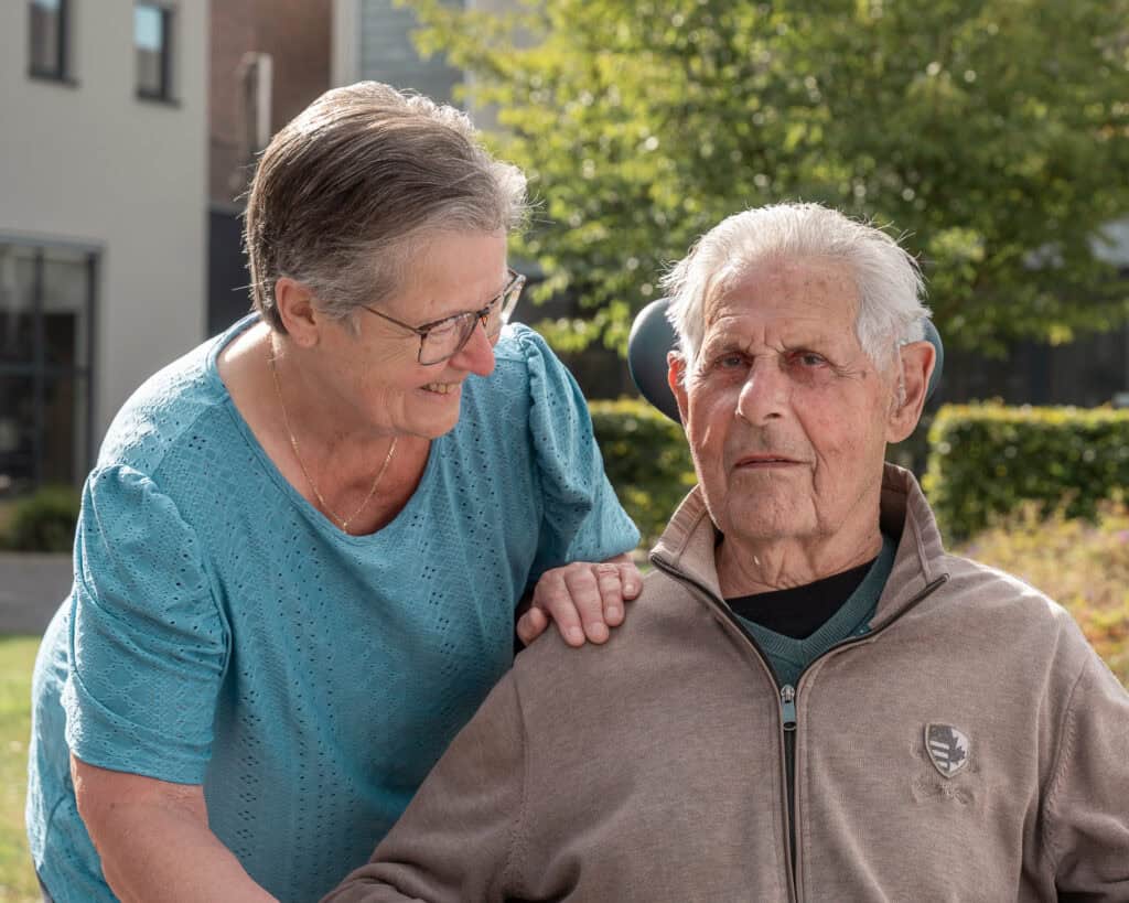 Een oudere man zit buiten in een rolstoel terwijl een oudere vrouw naast hem staat, glimlacht en haar hand op zijn schouder legt.