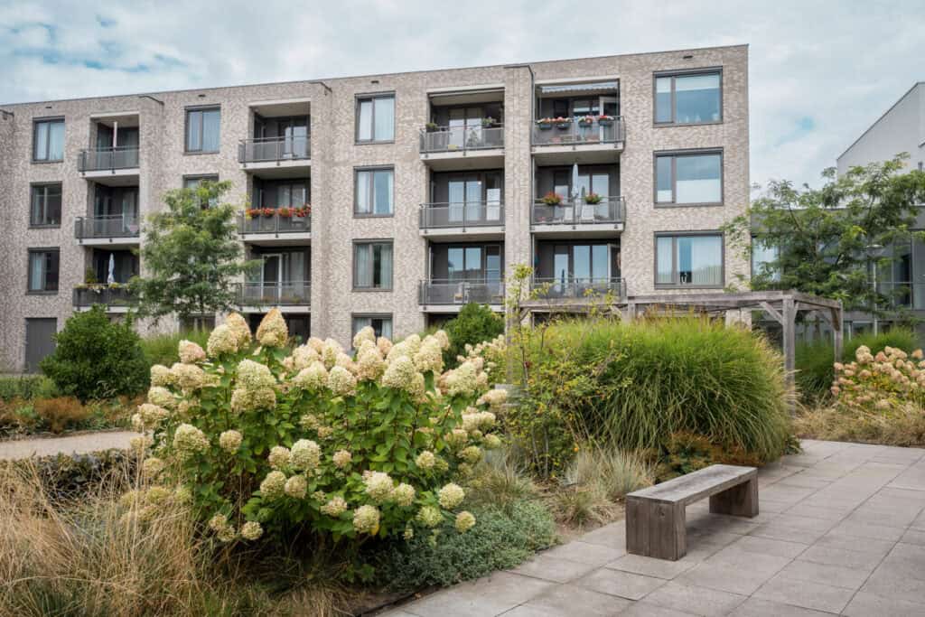 Een modern appartementengebouw met balkons kijkt uit op een aangelegde tuin met bloeiende struiken, groen en een houten bank op een geplaveid voetpad.
