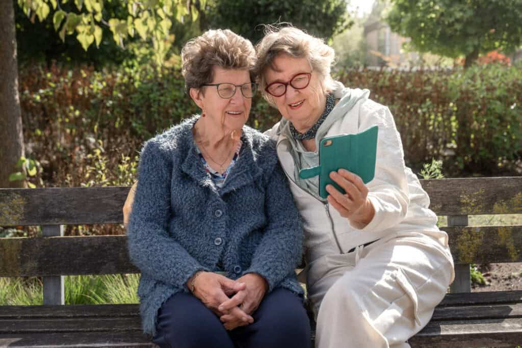 Gezellige senioren vrouwen kijken op smartphone op bank in park, ontspanning en sociale verbinding.